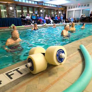 Seniors participating in a water aerobics class at an indoor pool, using foam dumbbells and noodles for exercise.
