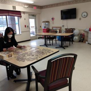 A person wearing a mask works on a puzzle at a table in a communal room.