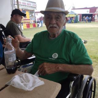An elderly man in a green shirt and hat enjoying a meal outdoors while seated in a wheelchair.