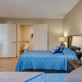 A cozy bedroom with two double beds, featuring blue bedding and warm lighting from table lamps. A doorway leads to an adjoining bathroom.