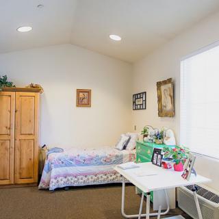 A cozy and well-decorated bedroom with a single bed, wooden wardrobe, and a small table adorned with pictures and knick-knacks.