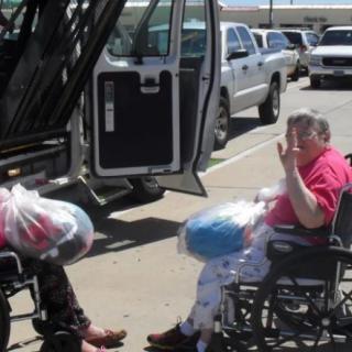 Two people in wheelchairs with shopping bags near a vehicle in a parking lot.