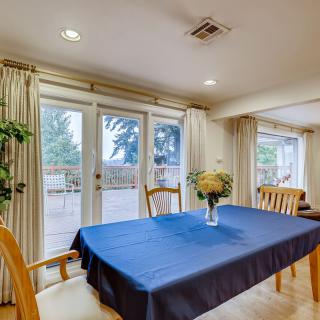 A cozy dining room featuring a table with a blue tablecloth, surrounded by wooden chairs and adorned with a vase of flowers. Natural light streams in through the sliding glass doors framed by curtains.