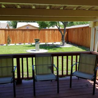 A cozy backyard view from a covered deck with three chairs facing a green lawn and wooden fence.
