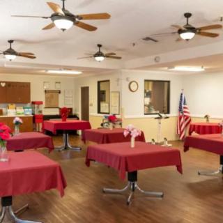 A spacious indoor dining area with tables covered in red tablecloths, wooden flooring, and ceiling fans.