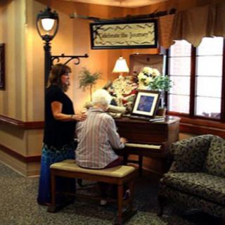 An elderly person playing the piano with a caregiver standing nearby in a cozy indoor setting.