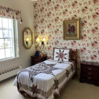 A cozy bedroom with floral wallpaper and a single bed adorned with a quilt, featuring traditional decor elements like a framed artwork and mirror.
