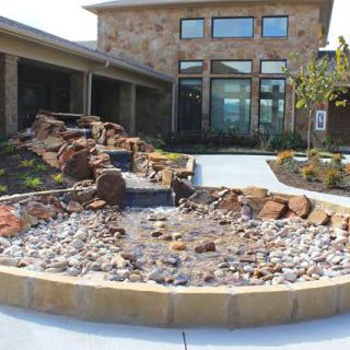 A stone building with large windows features a landscaped garden with a water feature and rocks.