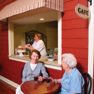 Two seniors enjoy a conversation over drinks at a cozy cafe, with a staff member serving from the counter.