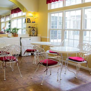 A bright cafe interior with white tables and chairs, accented by red cushions, and large windows letting in natural light.