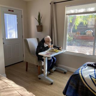 An elderly person enjoys a meal by the window in a sunny room.