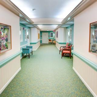 A well-lit hallway with carpeted flooring, featuring chairs and paintings on the walls.