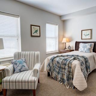 A cozy bedroom featuring a single bed, striped armchair, and soft lighting from a bedside lamp.