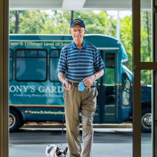 An elderly man in a striped shirt and hat walks with his dog near a bus.