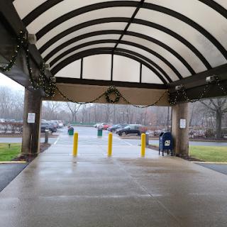 A covered entrance leading to a parking area, featuring yellow barriers and a view of the surrounding trees under overcast skies.