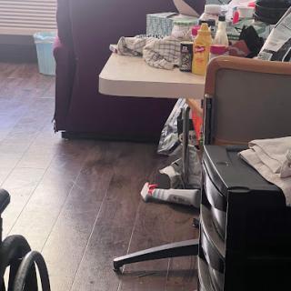 A cluttered sitting area featuring a table, wheelchair, and various personal items.
