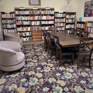 A cozy library room with bookshelves, a large table, and comfortable chairs on a colorful carpet.