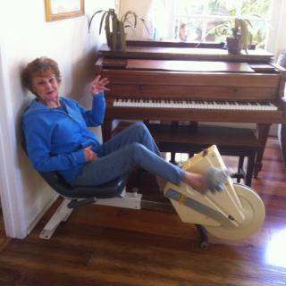A person is using a stationary exercise bike positioned next to a piano indoors.