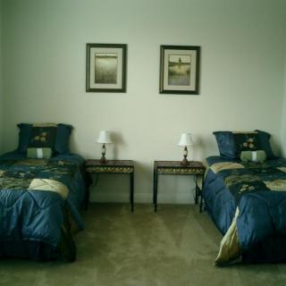 A neatly arranged bedroom with twin beds and matching bedding, flanked by bedside tables and lamps, adorned with framed art on the wall.