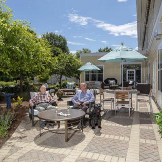 Two people sitting on a sunny patio with chairs and umbrellas, surrounded by greenery.