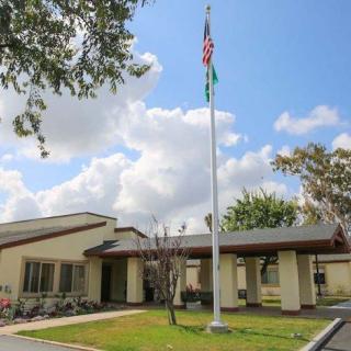 A single-story building with a flagpole in front, set against a backdrop of trees and a partly cloudy sky.