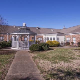 A scenic view of a brick building with a gazebo and pathway surrounded by shrubs and a lawn under a clear blue sky.