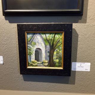 A charming painting of a house surrounded by lush trees, elegantly framed and displayed in an art gallery.