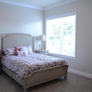 A cozy bedroom featuring a bed with floral bedding, a bedside table with a lamp, and a large window providing natural light.