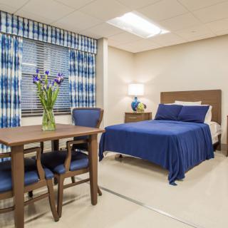 A neatly decorated bedroom featuring a blue color scheme, with a bed, table and chairs, and flowers in a vase by the window.