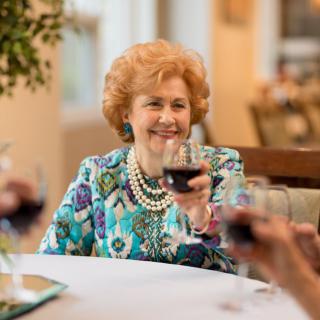 An elderly woman enjoying a social gathering with friends at a dining table, raising a glass of wine in celebration.