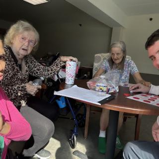 A group of people, including elderly individuals, gathered around a table engaging in conversation and activities.