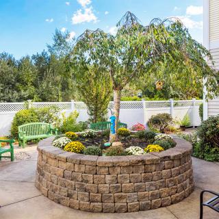 A beautifully landscaped patio with a circular stone planter featuring lush greenery and trees, set against a bright, clear day.