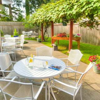 A sunny garden patio with white tables and chairs, surrounded by greenery.