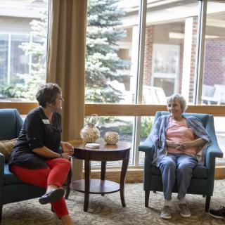 Two women engaged in a conversation in a comfortable indoor setting, possibly a senior living facility.