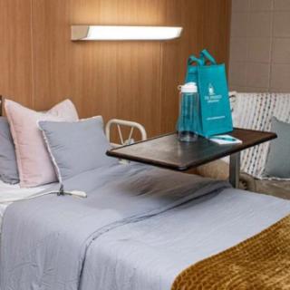 A hospital room featuring a bed with neatly arranged pillows, a blanket, and a tray table with a gift bag on top.