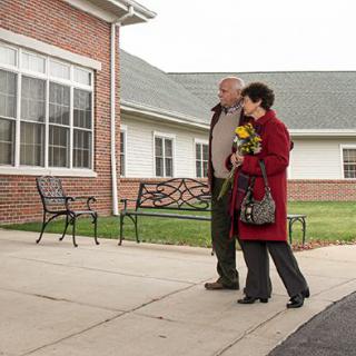 An elderly couple walking outside, carrying flowers near a building.
