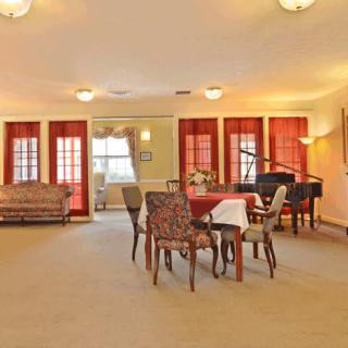 A spacious living room with a dining area and piano, featuring carpeted flooring and large windows with red curtains.