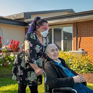 A caregiver assists an elderly person in a wheelchair, surrounded by blooming flowers on a sunny day.