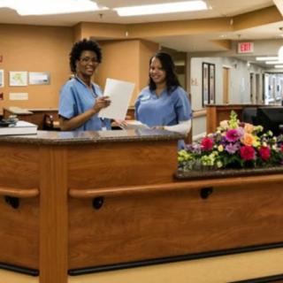 Two nurses stand at the hospital reception desk, smiling and holding papers.