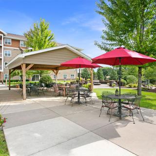 A serene outdoor patio area with tables and chairs under red umbrellas, adjacent to a gazebo and surrounded by lush greenery.