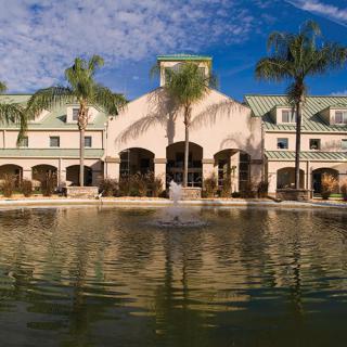 A scenic view of a building surrounded by palm trees, with a fountain reflecting in the water under a partly cloudy sky.
