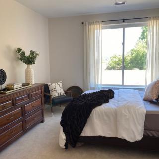 A minimalist bedroom with a large window allowing natural light, featuring a neatly made bed, a wooden dresser, and simple decor.