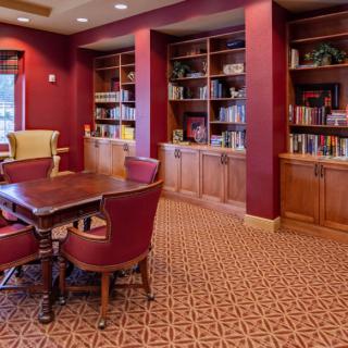 A cozy library room with red walls, bookshelves, and a central table surrounded by chairs.