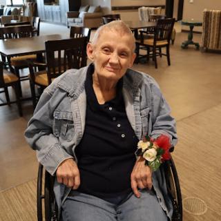 A smiling elderly woman in a wheelchair, adorned with a floral decoration, enjoying her time indoors.