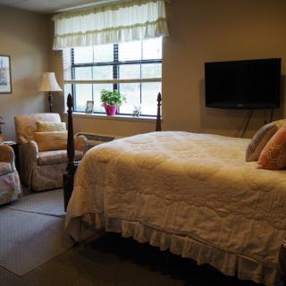 A cozy bedroom featuring a neatly made bed, an armchair by the window, and a wall-mounted television.