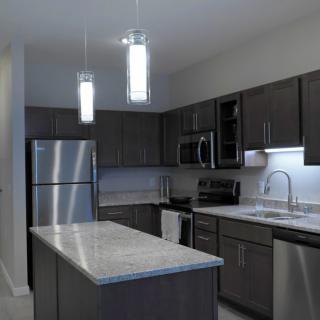 A modern kitchen with dark cabinets and stainless steel appliances features an island counter and elegant pendant lighting.