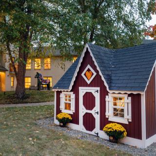 A quaint red playhouse sits in the yard, surrounded by trees and autumn decor, with a larger home visible in the background during an evening setting.