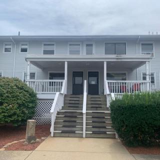 A two-story residential building with a porch and stairs leading to twin front doors, surrounded by bushes.