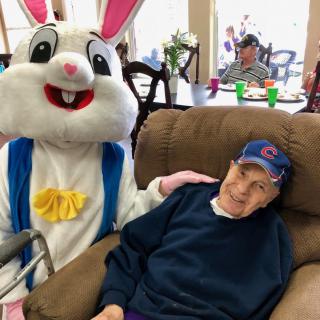 An Easter Bunny visits a nursing home, sharing a moment with an elderly resident in a recliner.