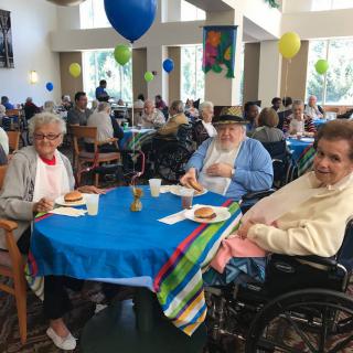 A group of seniors enjoying a community event inside a hall, adorned with colorful balloons and decorated tables.
